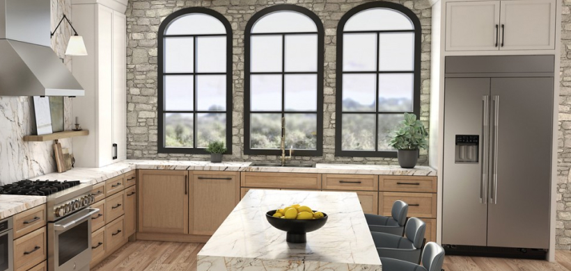 Une cuisine comprenant une cuisinière JennAir et une hotte JennAir, un four à micro-ondes JennAir et un réfrigérateur JennAir. On voit un îlot avec trois chaises et un bol de citrons. Sur le comptoir et les étagères se trouvent des planches à découper, des plantes et plusieurs autres objets.